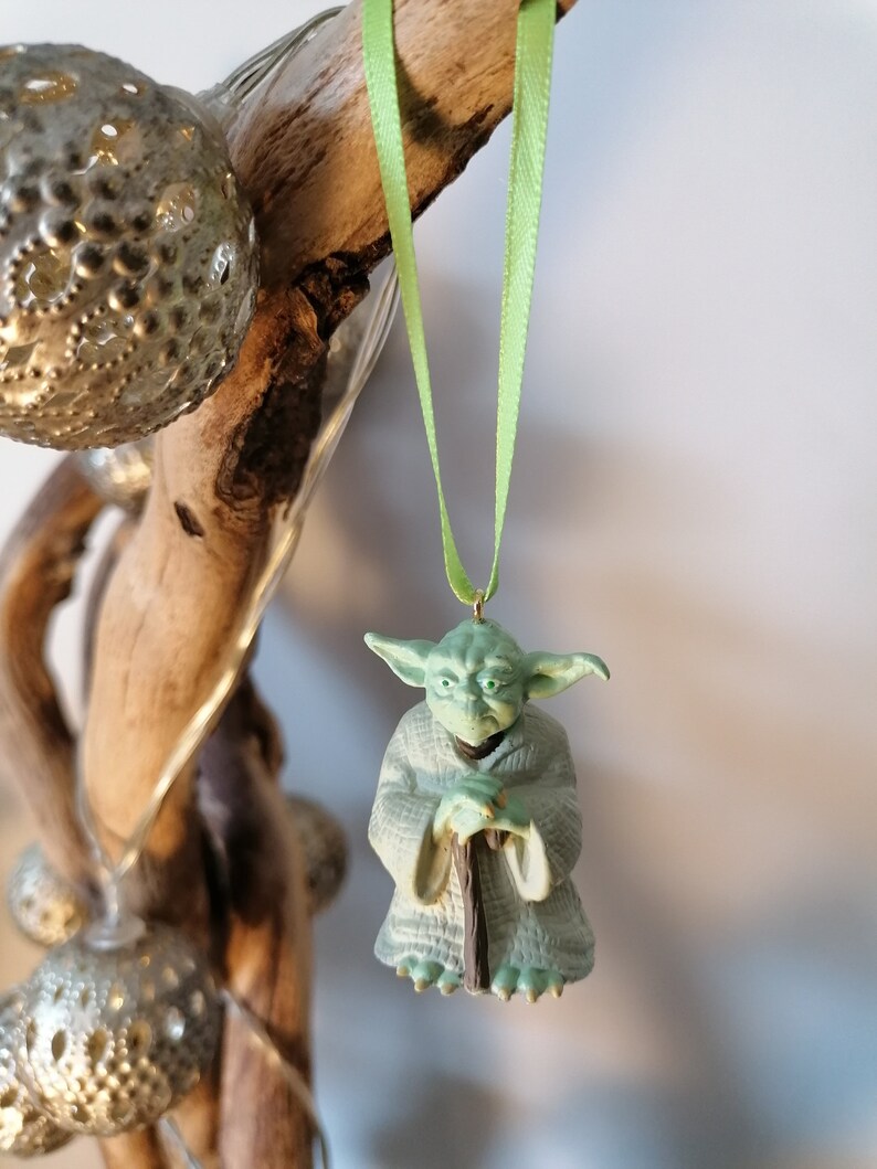 May include: A green Yoda ornament hanging from a light green ribbon. The ornament is made of plastic and features Yoda holding a staff.