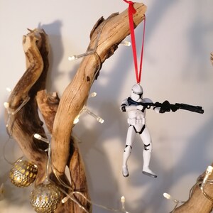 Puede incluir: Un adorno de figura de acci&oacute;n de Stormtrooper blanco colgado de una rama con una cinta roja. La figura sostiene un rifle bl&aacute;ster negro y est&aacute; en posici&oacute;n de disparo.