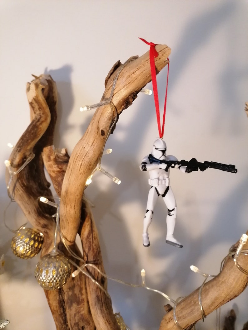 Puede incluir: Un adorno de figura de acci&oacute;n de Stormtrooper blanco colgado de una rama con una cinta roja. La figura sostiene un rifle bl&aacute;ster negro y est&aacute; en posici&oacute;n de disparo.