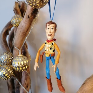 Puede incluir: Una figura de juguete de pl&aacute;stico de Woody de Toy Story colgando de una cinta azul. Woody lleva su caracter&iacute;stico sombrero de vaquero, chaleco y pantalones vaqueros. Sonr&iacute;e y tiene una expresi&oacute;n amigable.