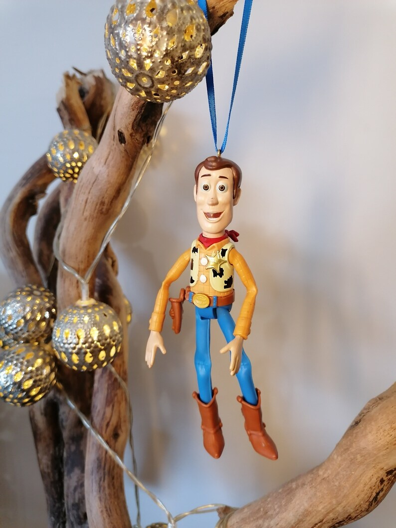 Puede incluir: Una figura de juguete de pl&aacute;stico de Woody de Toy Story colgando de una cinta azul. Woody lleva su caracter&iacute;stico sombrero de vaquero, chaleco y pantalones vaqueros. Sonr&iacute;e y tiene una expresi&oacute;n amigable.