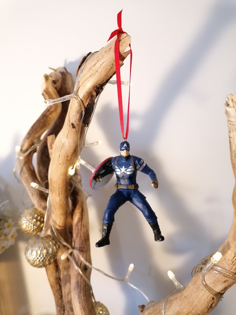 May include: A red ribbon hangs from a branch of driftwood, with a Captain America action figure ornament attached. The figure is in a flying pose, holding a shield.