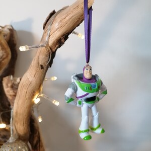 May include: A green and white plastic toy figurine of Buzz Lightyear from the Toy Story movie franchise. The figurine is hanging from a purple ribbon and is a Christmas ornament.