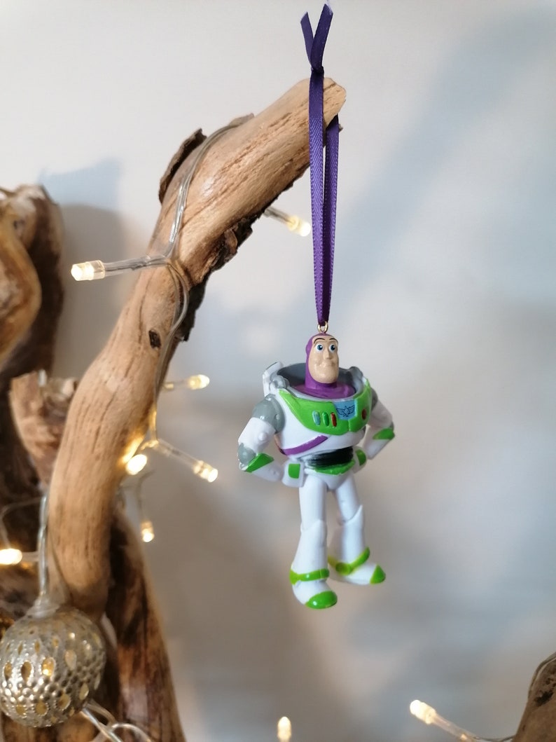 May include: A green and white plastic toy figurine of Buzz Lightyear from the Toy Story movie franchise. The figurine is hanging from a purple ribbon and is a Christmas ornament.