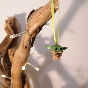 May include: A green and brown figurine of a character from the Star Wars franchise, hanging from a green ribbon on a branch of driftwood. The figurine is holding a small cup.