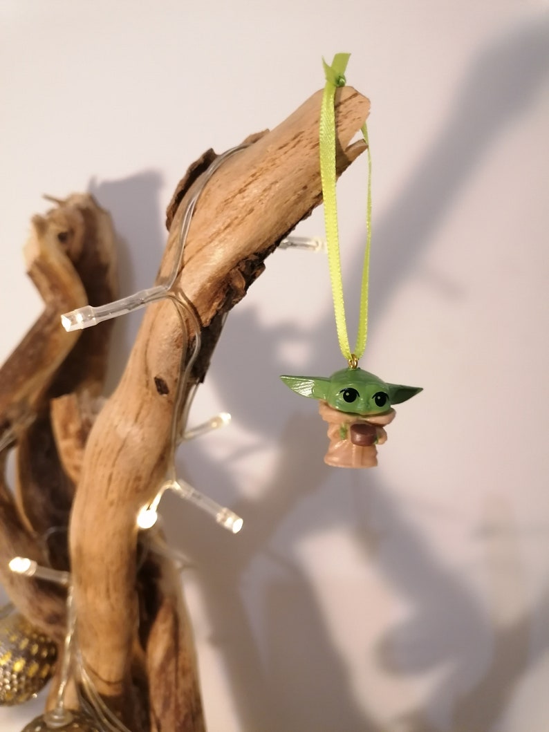 May include: A green and brown figurine of a character from the Star Wars franchise, hanging from a green ribbon on a branch of driftwood. The figurine is holding a small cup.