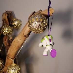 Puede incluir: Una figura de juguete de astronauta blanco y verde colgando de una rama con una cinta morada. El astronauta lleva un casco morado y tiene un pie morado. La rama est&aacute; decorada con adornos dorados y blancos.