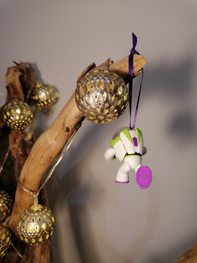 Puede incluir: Una figura de juguete de astronauta blanco y verde colgando de una rama con una cinta morada. El astronauta lleva un casco morado y tiene un pie morado. La rama est&aacute; decorada con adornos dorados y blancos.
