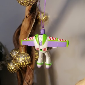 Peut inclure: Ornement de No&euml;l Buzz l'&Eacute;clair, avec un corps blanc, des d&eacute;tails verts et des ailes violettes. Le soldat jouet est suspendu par un ruban violet. Des guirlandes lumineuses dor&eacute;es sont en arri&egrave;re-plan.