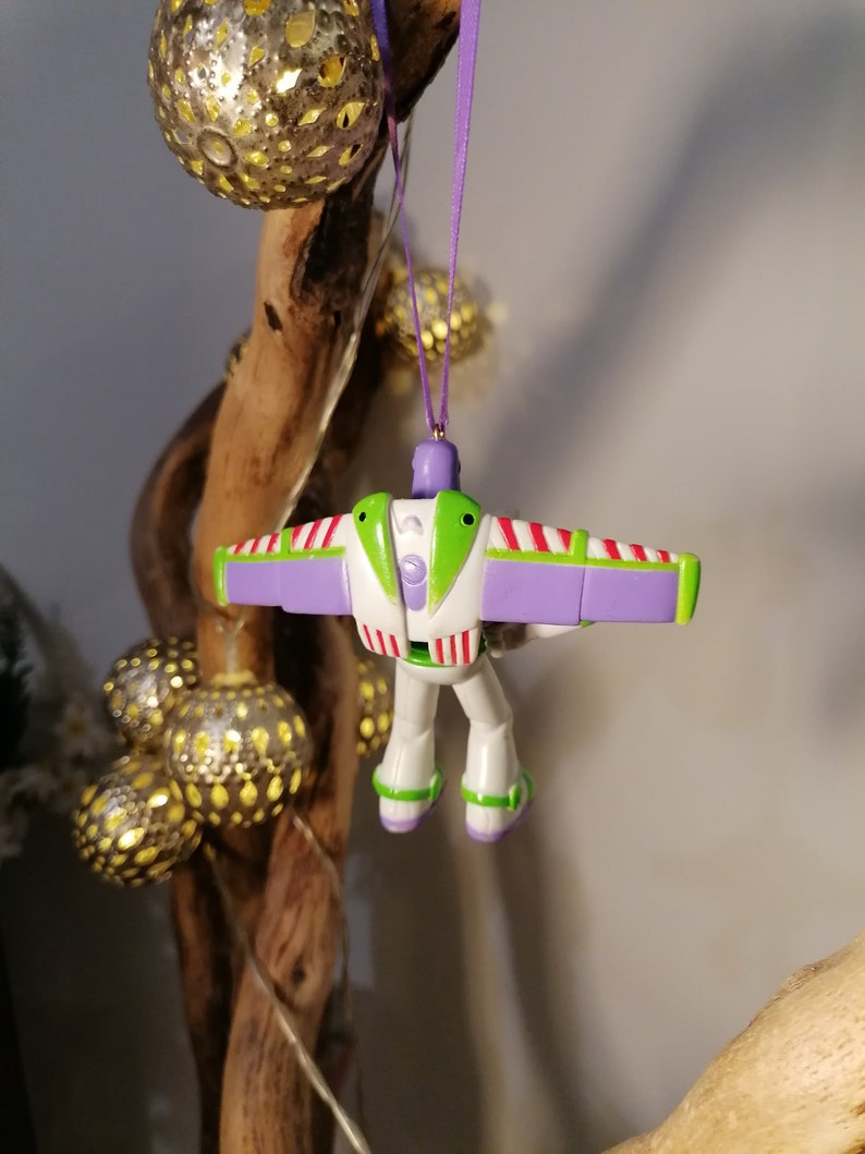 Peut inclure: Ornement de No&euml;l Buzz l'&Eacute;clair, avec un corps blanc, des d&eacute;tails verts et des ailes violettes. Le soldat jouet est suspendu par un ruban violet. Des guirlandes lumineuses dor&eacute;es sont en arri&egrave;re-plan.