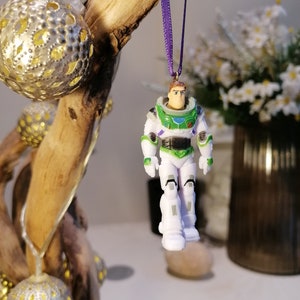 May include: A white and green plastic toy astronaut ornament with a purple ribbon. The astronaut is wearing a space suit with a helmet and has a friendly face.