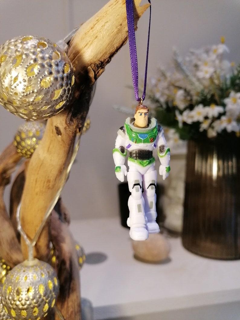 May include: A white and green plastic toy astronaut ornament with a purple ribbon. The astronaut is wearing a space suit with a helmet and has a friendly face.