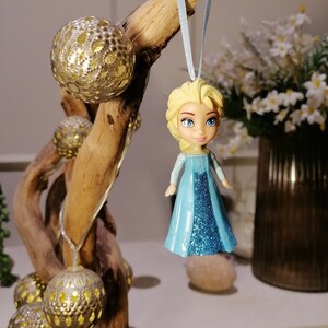 May include: A blue and silver glitter bauble featuring a small plastic doll with blonde hair and a blue dress. The bauble is hanging from a blue ribbon on a driftwood branch.
