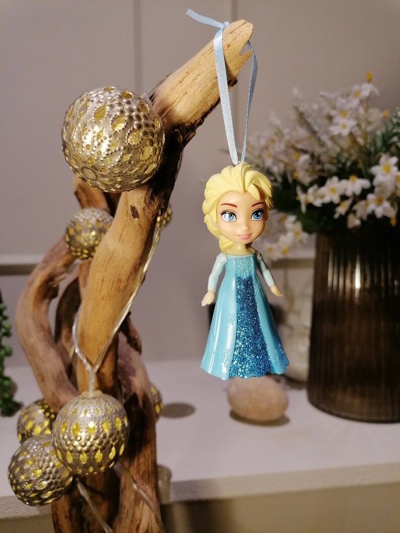 May include: A blue and silver glitter bauble featuring a small plastic doll with blonde hair and a blue dress. The bauble is hanging from a blue ribbon on a driftwood branch.