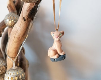 May include: A small, beige lion cub figurine with green eyes, sitting on a gray base. The figurine is hanging from a gold ribbon.
