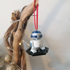 May include: A white and blue R2-D2 Star Wars character ornament hanging from a branch with a red ribbon. The ornament is on a black base.