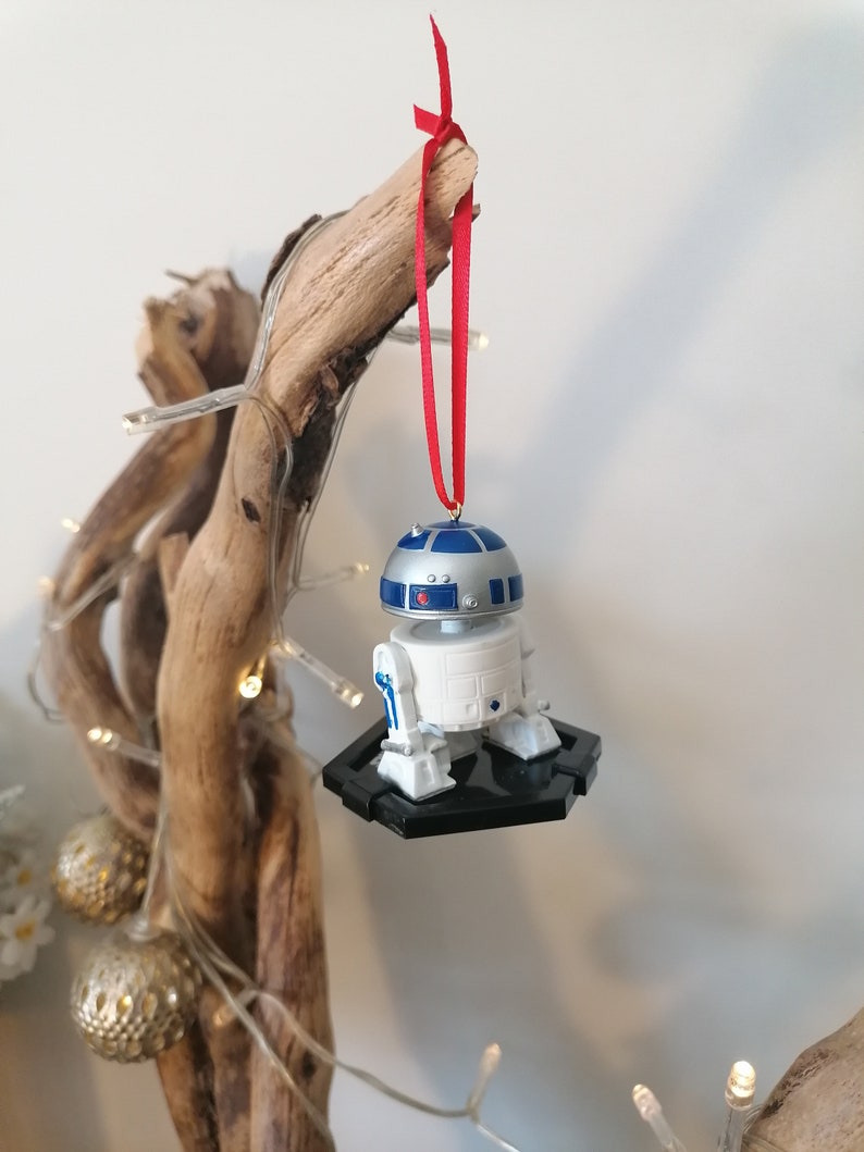 May include: A white and blue R2-D2 Star Wars character ornament hanging from a branch with a red ribbon. The ornament is on a black base.