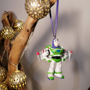Pu&ograve; includere: Un ornamento di astronauta di plastica bianco e verde con un nastro viola appeso a un ramo con luci a catena. L'astronauta indossa un casco blu e ha un razzo rosso e bianco attaccato alla schiena.