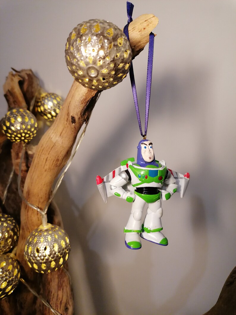 Pu&ograve; includere: Un ornamento di astronauta di plastica bianco e verde con un nastro viola appeso a un ramo con luci a catena. L'astronauta indossa un casco blu e ha un razzo rosso e bianco attaccato alla schiena.