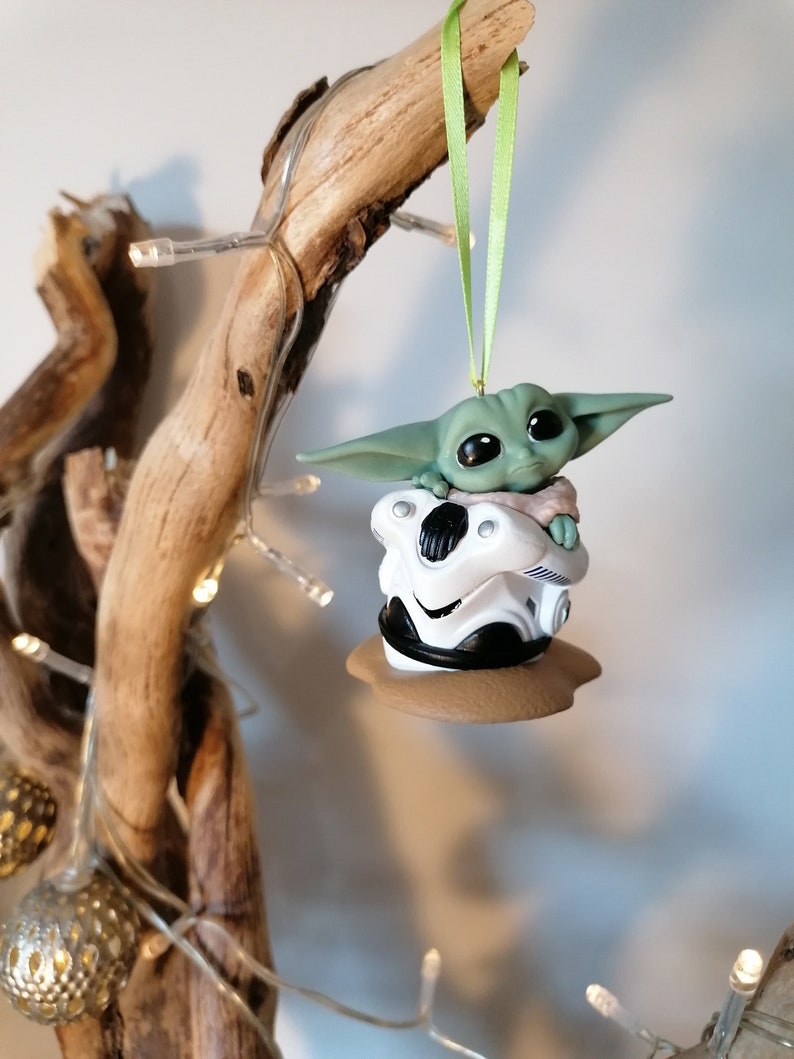 K&ouml;nnte beinhalten: Ein gr&uuml;ner und wei&szlig;er Weihnachtsbaumschmuck mit einer kleinen, gr&uuml;nen Alienfigur, die in einem wei&szlig;en und schwarzen Raumschiff sitzt. Der Schmuck h&auml;ngt an einem gr&uuml;nen Band an einem Zweig.