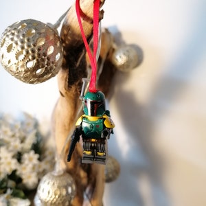 May include: A green and yellow Lego Star Wars Boba Fett ornament hanging from a piece of driftwood with a red ribbon.