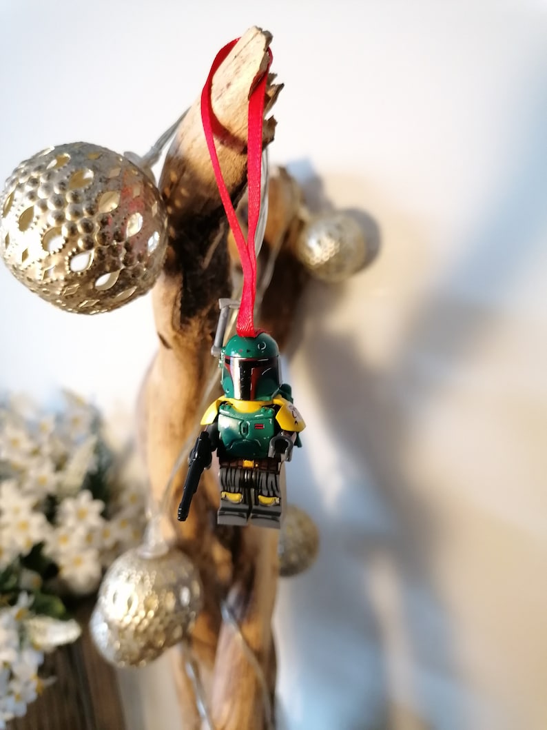 May include: A green and yellow Lego Star Wars Boba Fett ornament hanging from a piece of driftwood with a red ribbon.