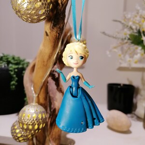 Peut inclure: Une figurine en plastique bleue d'une princesse aux cheveux blonds portant une robe bleue. La figurine est suspendue &agrave; un ruban bleu.