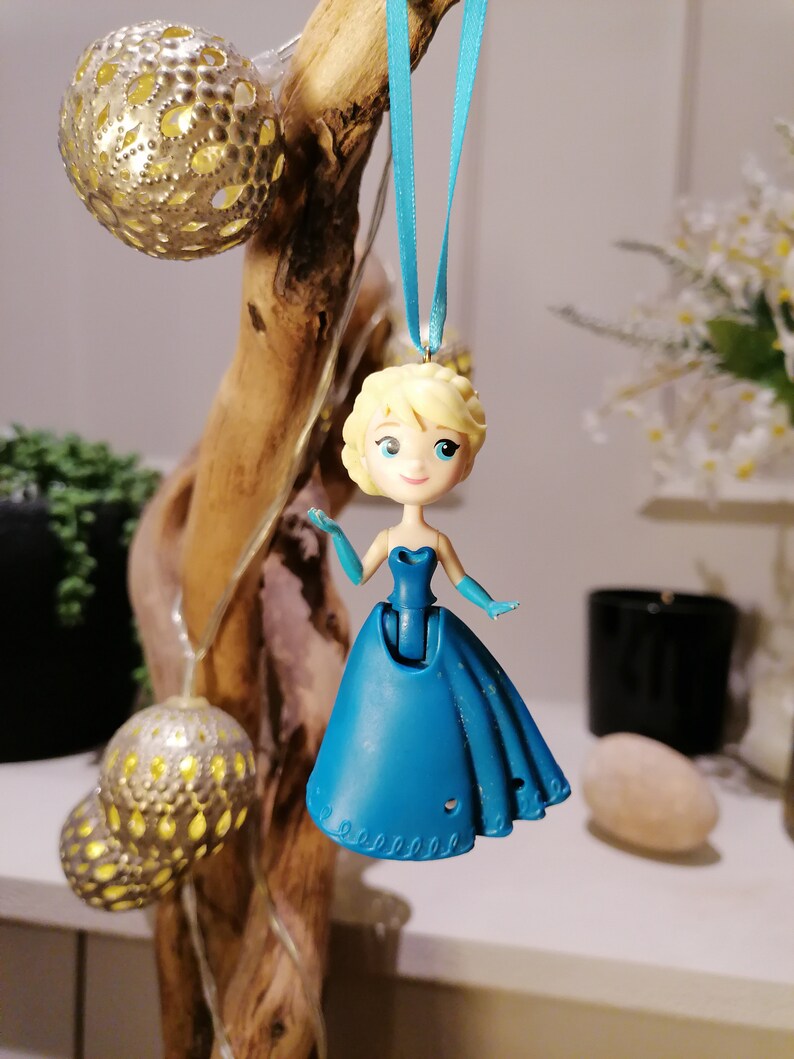 Peut inclure: Une figurine en plastique bleue d'une princesse aux cheveux blonds portant une robe bleue. La figurine est suspendue &agrave; un ruban bleu.