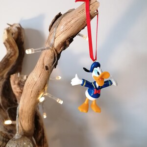 May include: A blue and yellow Donald Duck Christmas ornament hanging from a branch with a red ribbon. The ornament is made of plastic and has a 3D design.