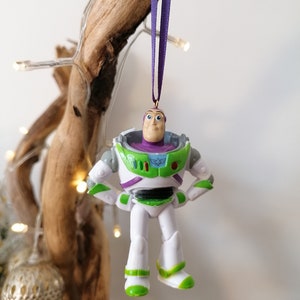 May include: A white and green plastic ornament of Buzz Lightyear from the Toy Story franchise. The ornament is hanging from a purple ribbon.