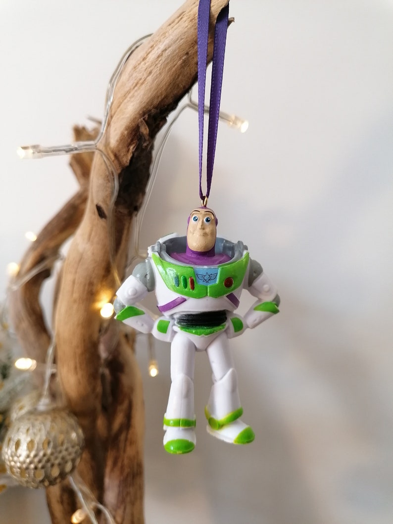 May include: A white and green plastic ornament of Buzz Lightyear from the Toy Story franchise. The ornament is hanging from a purple ribbon.