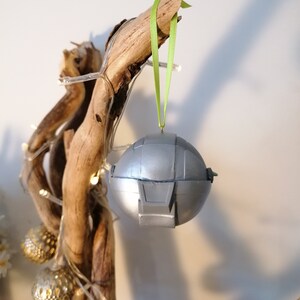 May include: A silver and gray Christmas ornament shaped like a spaceship, hanging from a green ribbon on a branch.