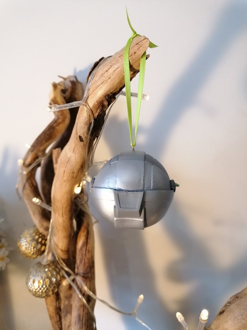 May include: A silver and gray Christmas ornament shaped like a spaceship, hanging from a green ribbon on a branch.