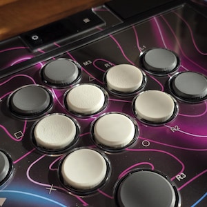 May include: A gaming controller interface featuring a black surface with pink and blue topographic lines. It includes multiple circular buttons in shades of white and dark gray, with labels such as R1, R2, R3, M3, and various geometric symbols.