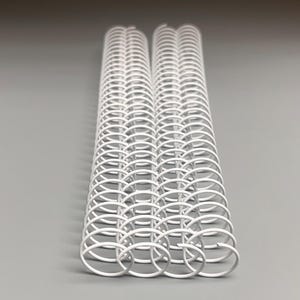 May include: Two stacks of white metal binding coils. The coils are made of a series of connected, rounded loops. The coils are used for binding documents, notebooks, or other paper products. The background is a solid, light gray color.