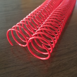 May include: Two red plastic binding coils, also known as coil binding or spiral binding, are lying on a wooden surface. The coils are interlocked and ready to be used for binding documents.