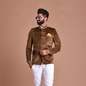 Puede incluir: Una chaqueta de terciopelo marrón con cuello mandarín, cierre de botones y un parche en el codo. La chaqueta tiene un bolsillo en el lado izquierdo y un pañuelo en el bolsillo derecho. El modelo lleva pantalones blancos y botas marrones.