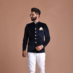 Puede incluir: Una chaqueta de terciopelo azul marino con cuello mandarín, botones dorados y un pañuelo de bolsillo blanco. La chaqueta se lleva sobre una camisa blanca y pantalones blancos.