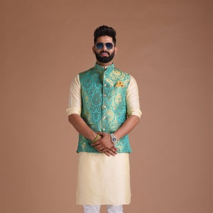 Puede incluir: Un hombre con un chaleco estampado en verde azulado y dorado sobre una camisa crema y pantalones blancos. Lleva gafas de sol y barba.