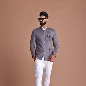 Puede incluir: Una chaqueta gris con cuello mandarín y cierre de botones. La chaqueta tiene dos bolsillos de solapa y un alfiler decorativo en el lado izquierdo. El modelo lleva pantalones blancos y zapatos marrones.