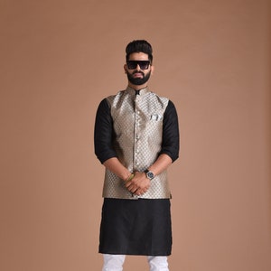 Puede incluir: Un hombre con un chaleco dorado estampado sobre una camisa negra de manga larga, combinado con una túnica negra, pantalones blancos y botas marrones. También lleva gafas de sol y un reloj.