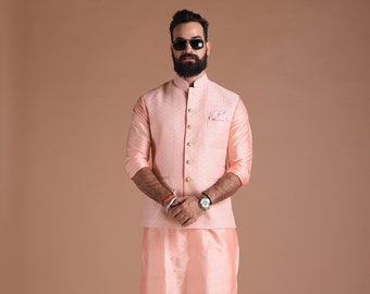 May include: A peach-colored kurta and matching waistcoat with gold buttons. The man is wearing white pants and brown shoes.
