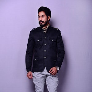 Puede incluir: Una chaqueta negra con cuatro bolsillos delanteros y botones plateados, combinada con pantalones grises claros y botas marrones. La chaqueta tiene charreteras. Se ve un pañuelo estampado.