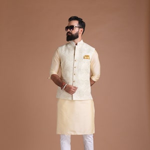 Puede incluir: Un hombre con una kurta y un chaleco a juego de color crema con bordados intrincados. Lleva pantalones blancos y zapatos marrones. También lleva gafas de sol.