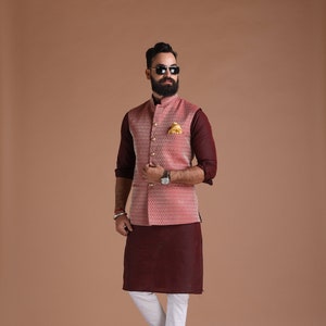 Puede incluir: Un hombre con una kurta marrón con un chaleco estampado rosa, pantalones blancos y zapatos negros. Lleva gafas de sol y barba.