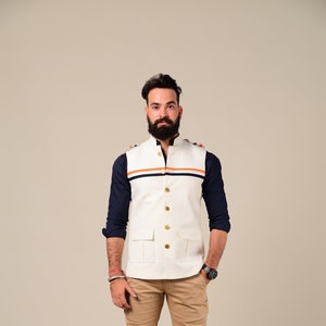 May include: A cream-colored vest with gold buttons and two front pockets, worn over a navy blue long-sleeve shirt. The vest has orange and navy blue stripes across the chest. The outfit is completed with khaki pants and brown leather boots.