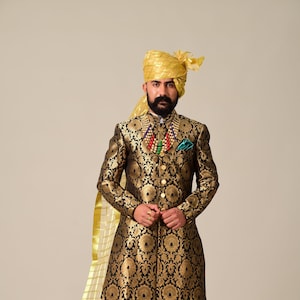 May include: A man wearing a black and gold patterned long-sleeved kurta with a gold turban, white pants, and brown shoes. He is wearing a beaded necklace with a green and red pendant.