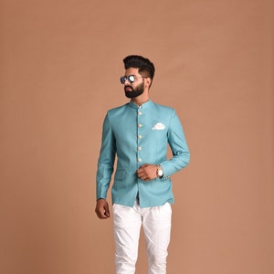 Puede incluir: Una chaqueta azul claro con botones dorados y un pañuelo de bolsillo blanco. La chaqueta se lleva sobre una camisa blanca con cuello y pantalones blancos.