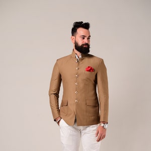 Puede incluir: Una chaqueta marrón con cuello mandarín y botones dorados. La chaqueta tiene un bolsillo a cada lado y un pañuelo de bolsillo rojo. La persona lleva pantalones blancos y zapatos marrones.