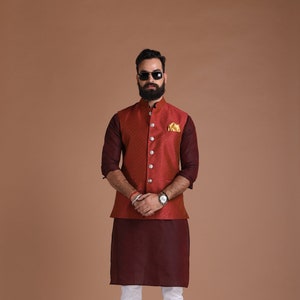 May include: A formal ensemble featuring a maroon kurta and white pants. A patterned, rust-colored vest with matching sleeves and a pocket square completes the look. The outfit is paired with black dress shoes and a watch.